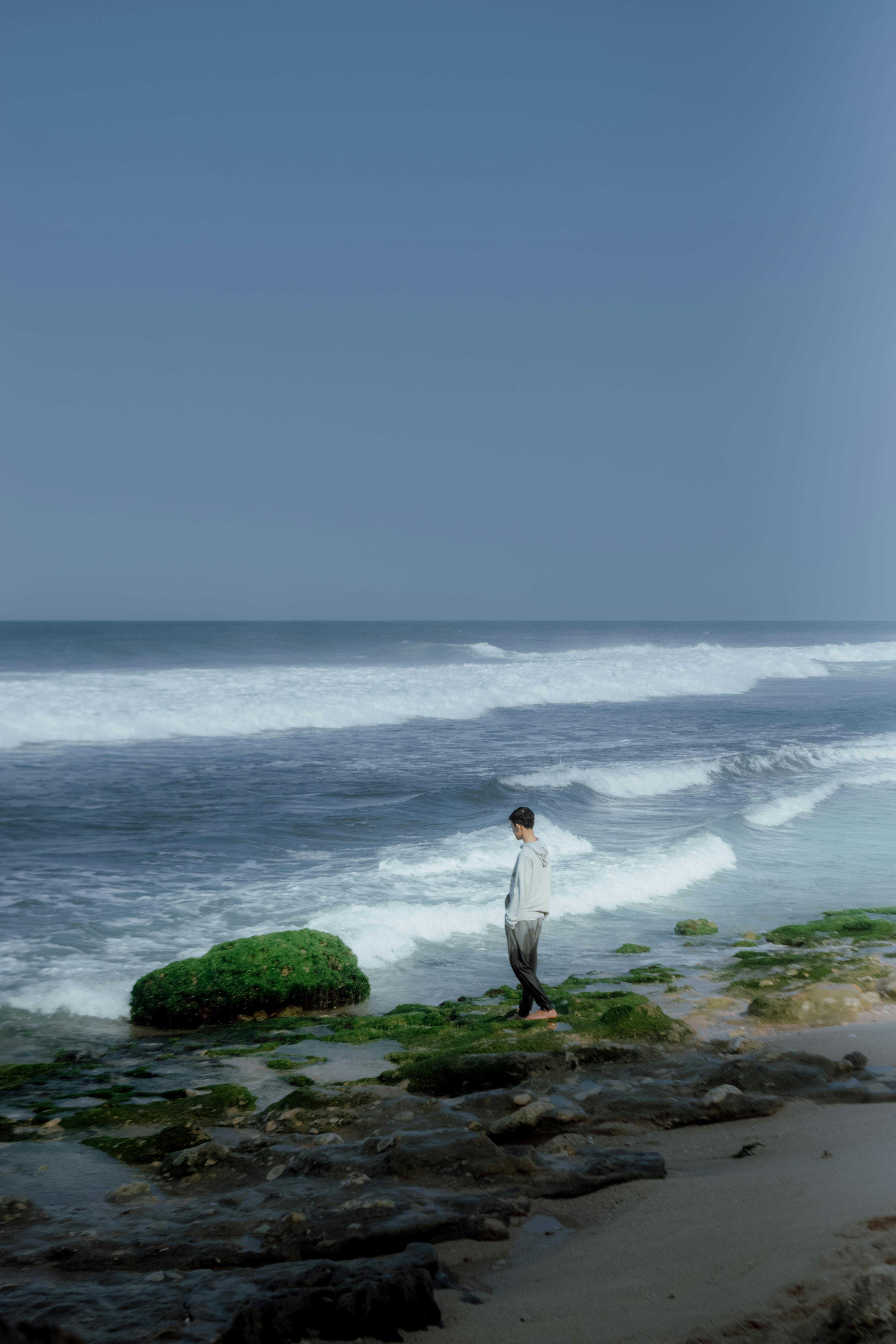 A person stands on moss-covered rocks by the ocean under a clear blue sky, surrounded by waves and seafoam.