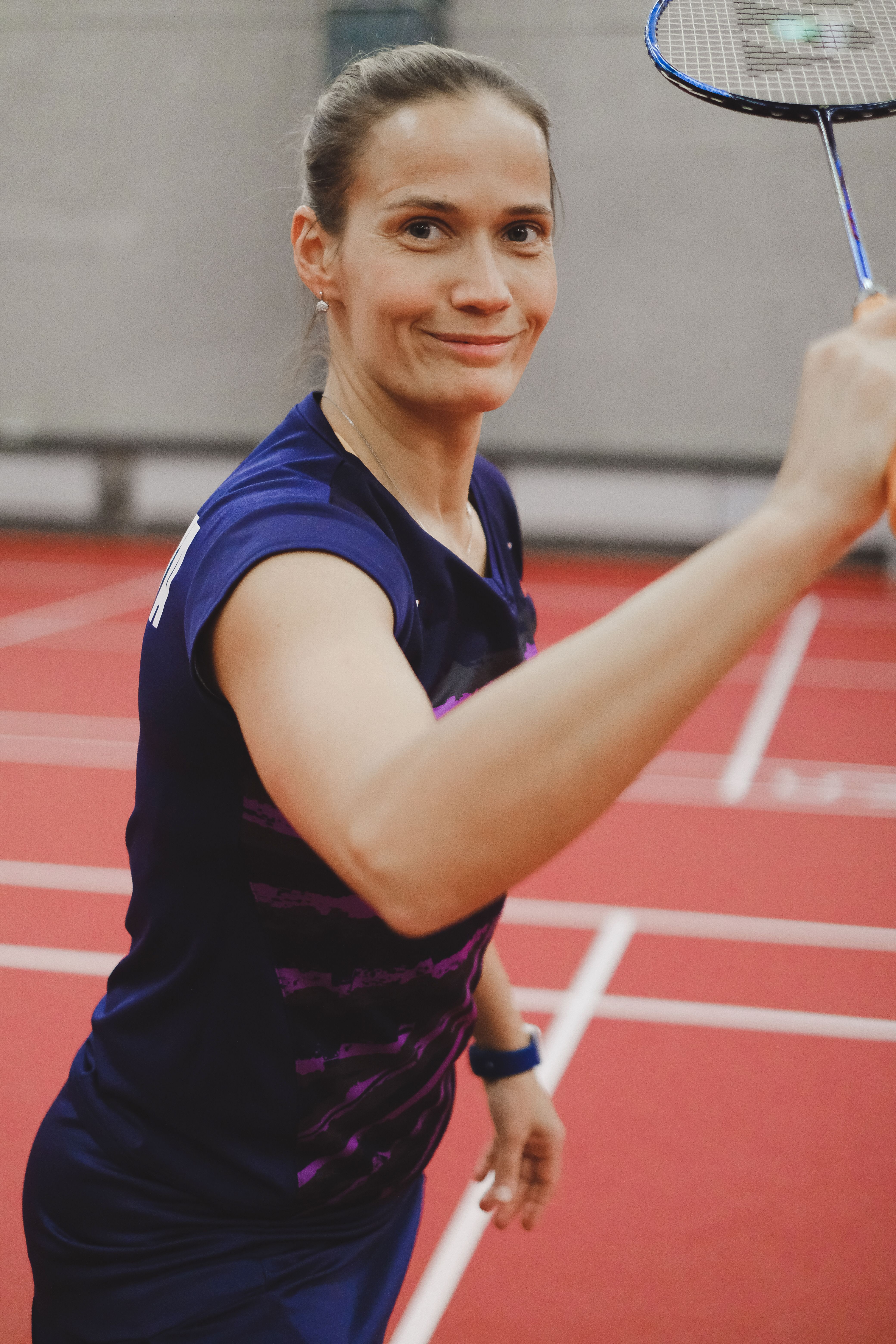 Portrait of a smiling woman playing badminton indoors on a red court.