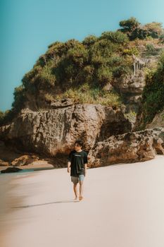 A man walks on a sandy beach with rocky cliffs under a clear blue sky.