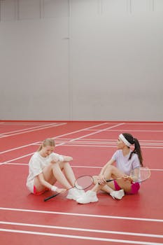Two young women sitting on a badminton court taking a break from playing.