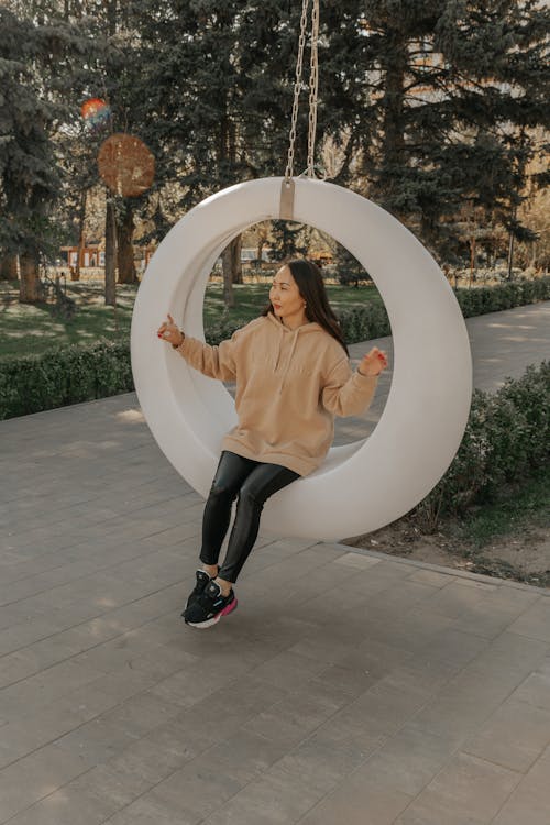 Free Cool young ethnic female in hoodie and sneakers sitting on ring swing while looking away in urban park Stock Photo