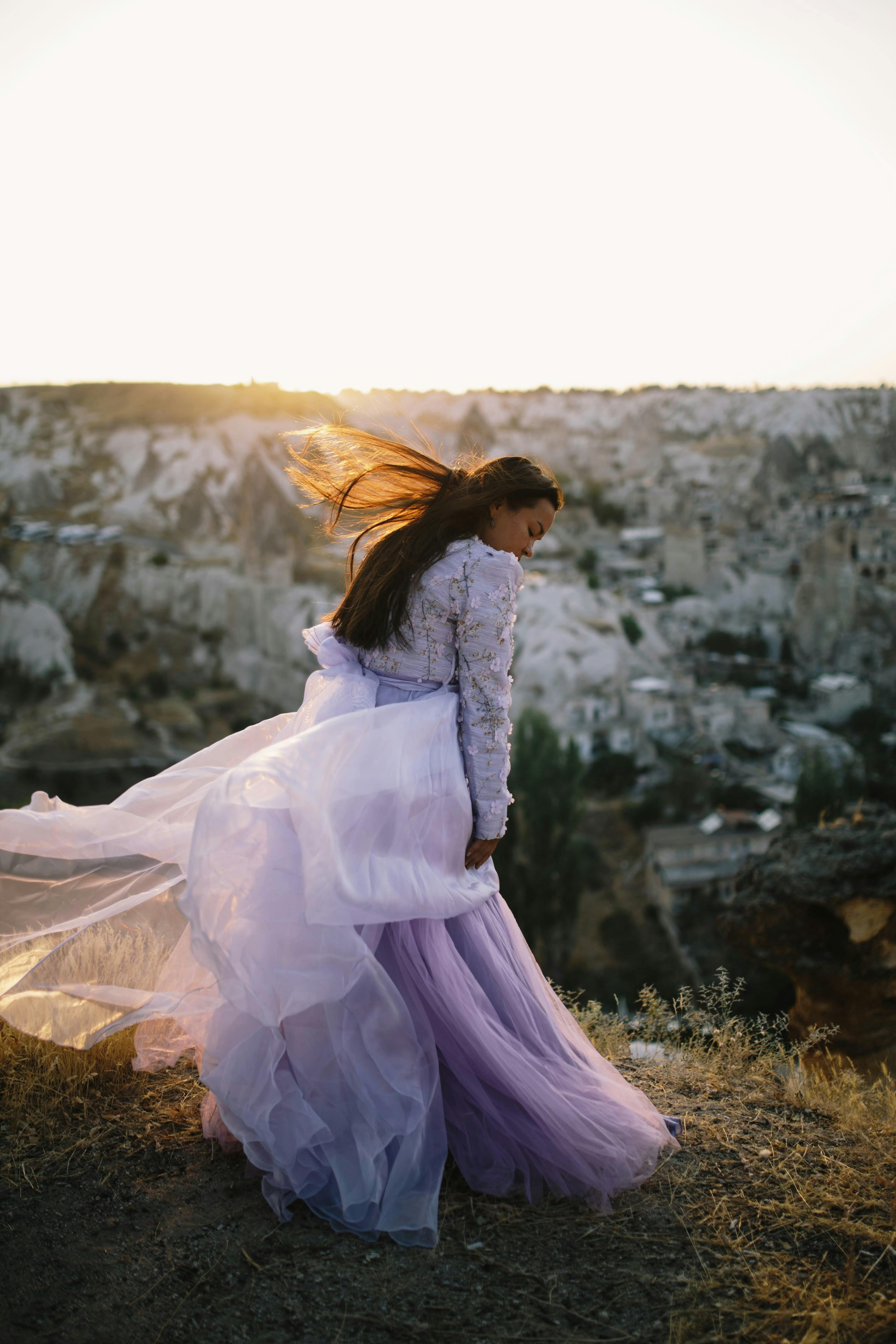 A woman in a purple dress stands on a cliffside with the wind blowing at sunset, capturing elegance and freedom.