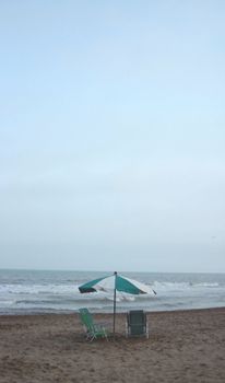 A peaceful beach setting featuring a lone umbrella and chairs on the sandy shore with waves in the background.