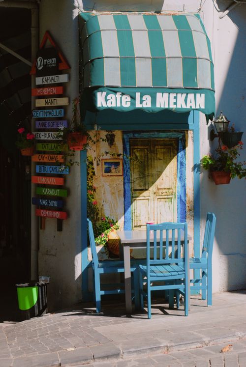 Free Sunlit alleyway cafe with blue chairs and vibrant decor. Ideal for travel themes. Stock Photo