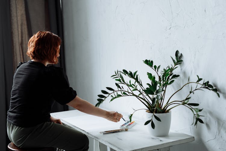 A Person Painting On A Desk Beside A Potted Plant