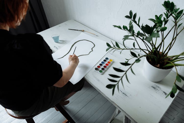 A Person Painting On A Desk Beside A Potted Plant