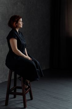 Side view of a woman in a black dress sitting indoors on a stool with dramatic lighting.
