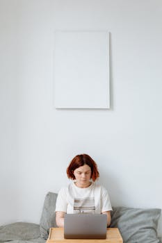 A woman sitting on a couch working remotely on a laptop at home with a blank frame above.