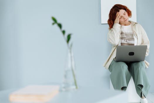 Woman sitting with laptop, holding head in a minimalist space, depicting stress and fatigue.