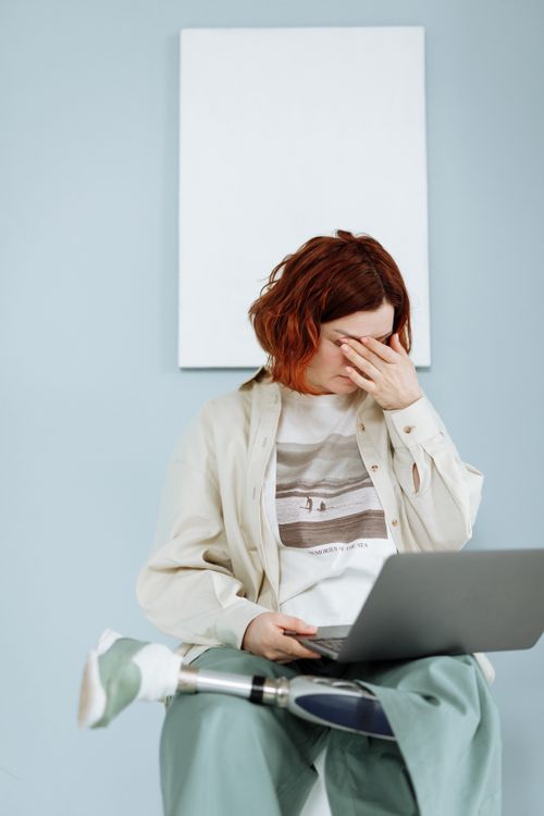 Free Woman with prosthetic leg sits indoors using a laptop, expressing frustration. Stock Photo