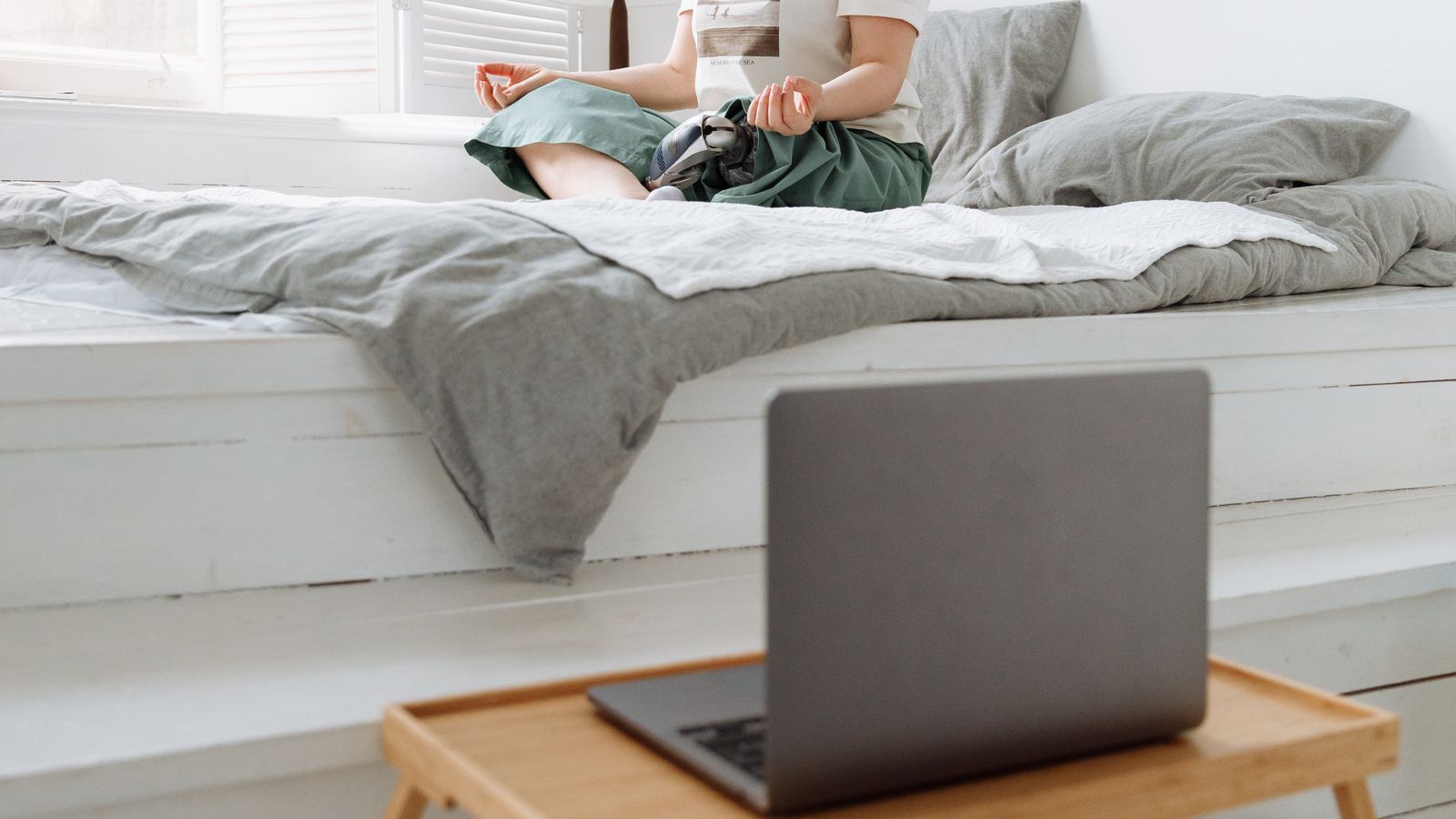 A woman meditating on a bed beside a bright window indoors