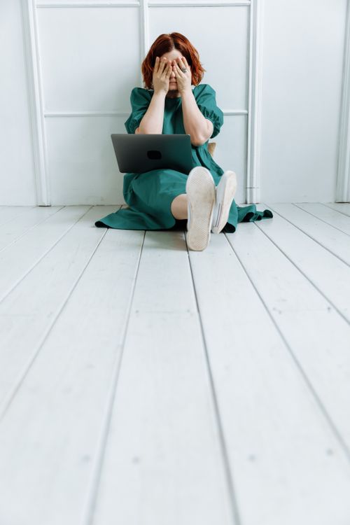 Free Woman in green dress sitting on floor with laptop, covering face in frustration. Stock Photo