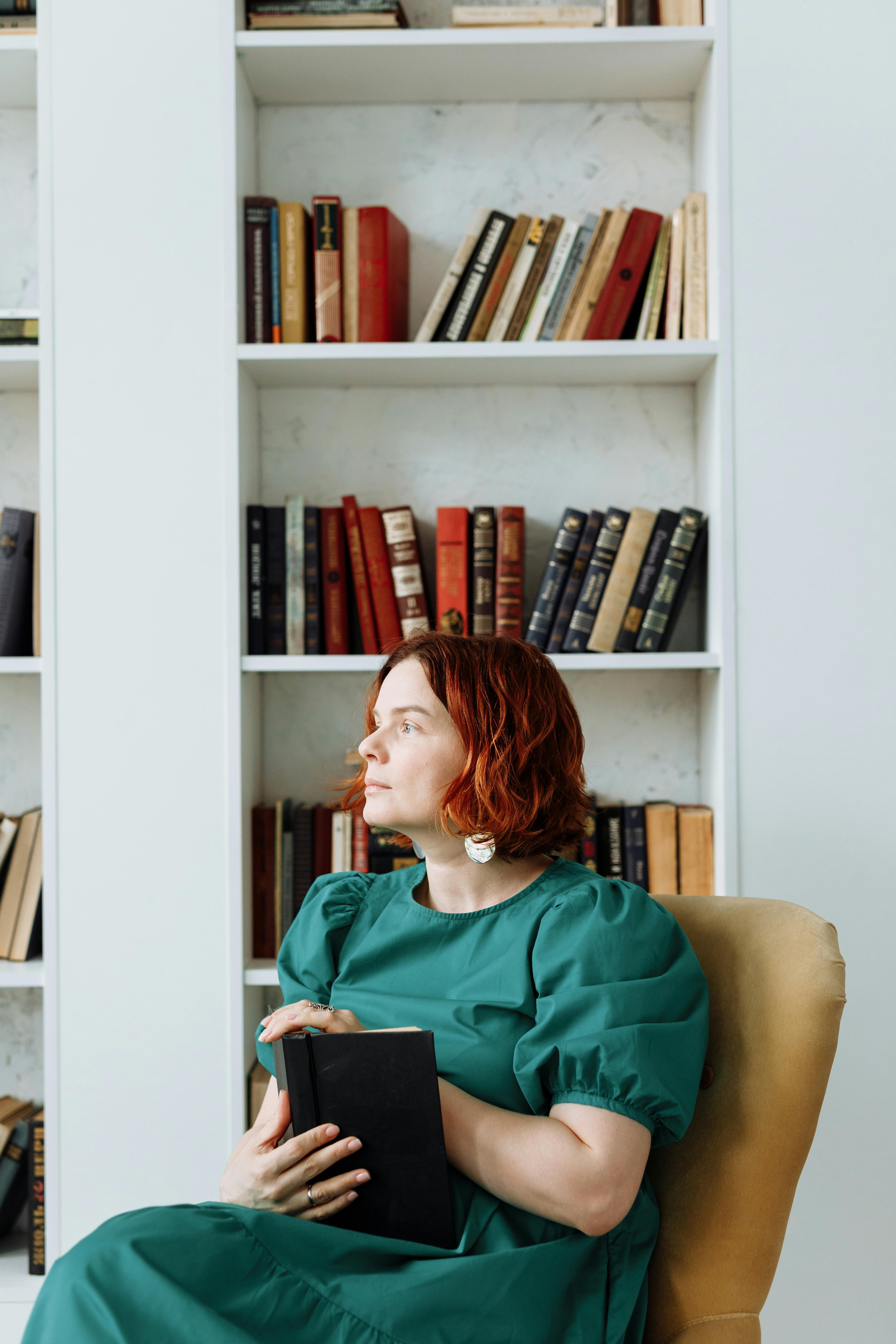 Pensive woman sitting in a library holding a book, surrounded by bookshelves.