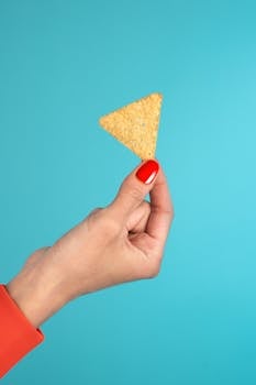 A hand with red nails holding a tortilla chip against a blue background.