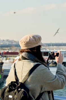 A woman with a backpack captures a scenic river view with birds using a camera during a winter day.