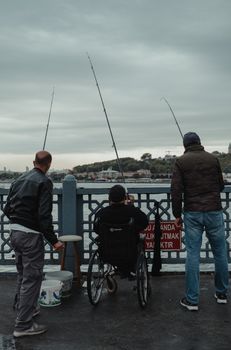 Three men fishing by the river, one in a wheelchair, under overcast skies.
