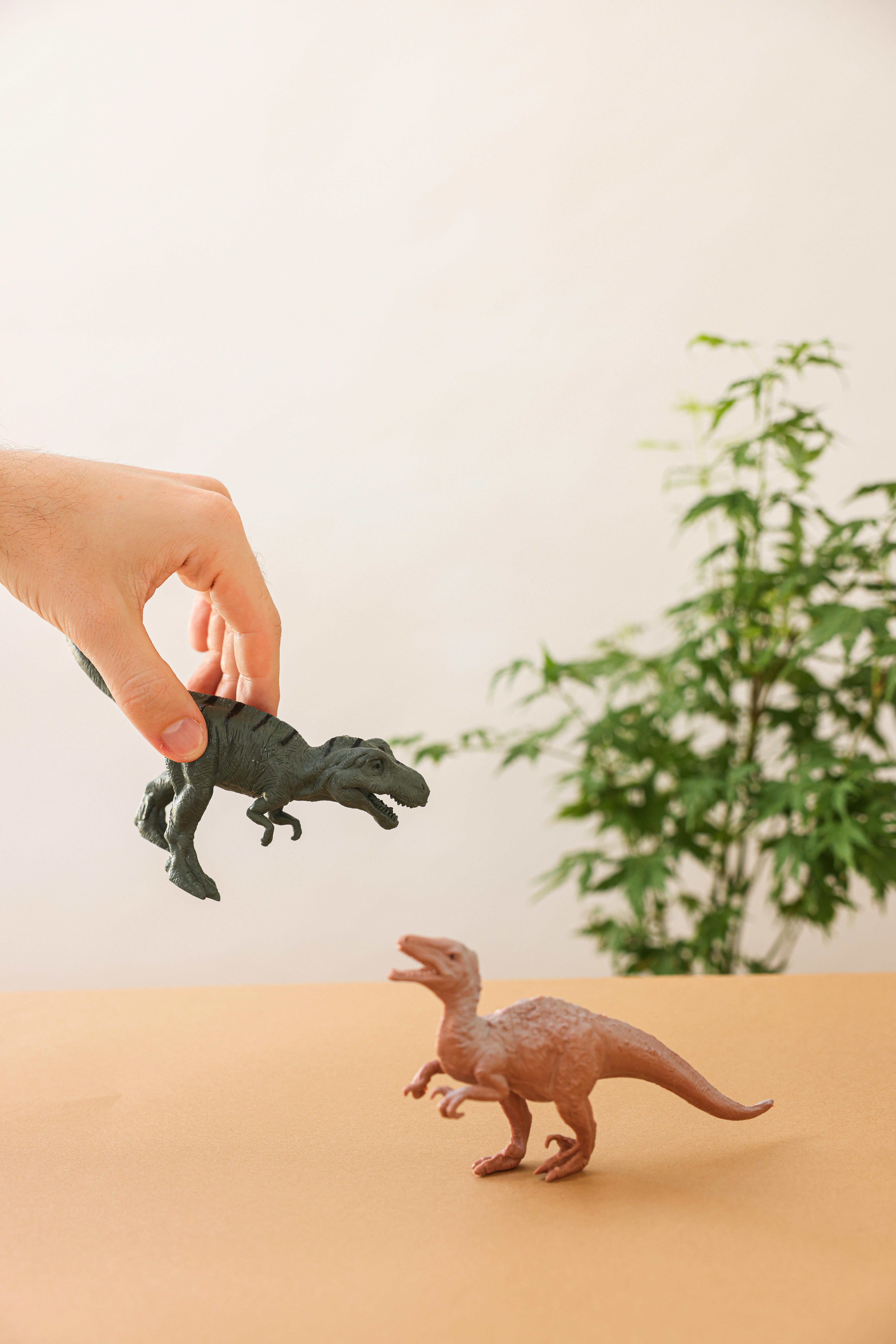 Hand interacting with dinosaur toys on a table, featuring green foliage background.