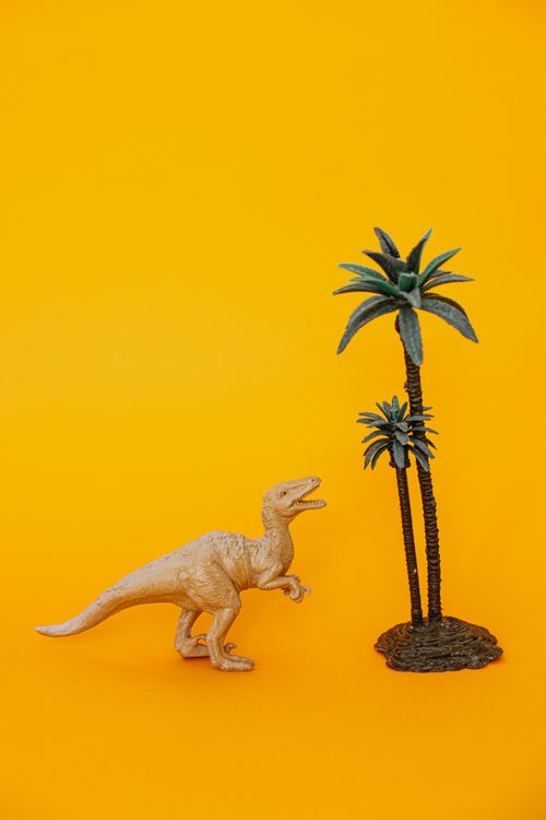Free Plastic dinosaur figurine next to palm tree on vibrant yellow backdrop. Stock Photo