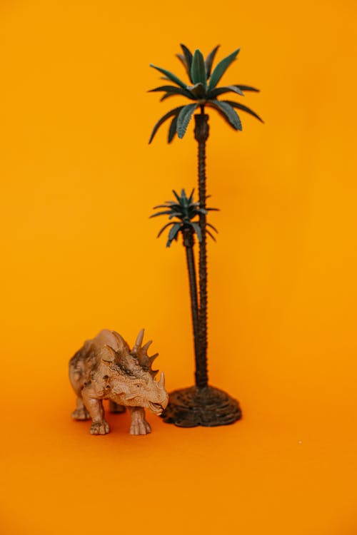 A Beige Toy Dinosaur Beside a Coconut Tree on Yellow Surface