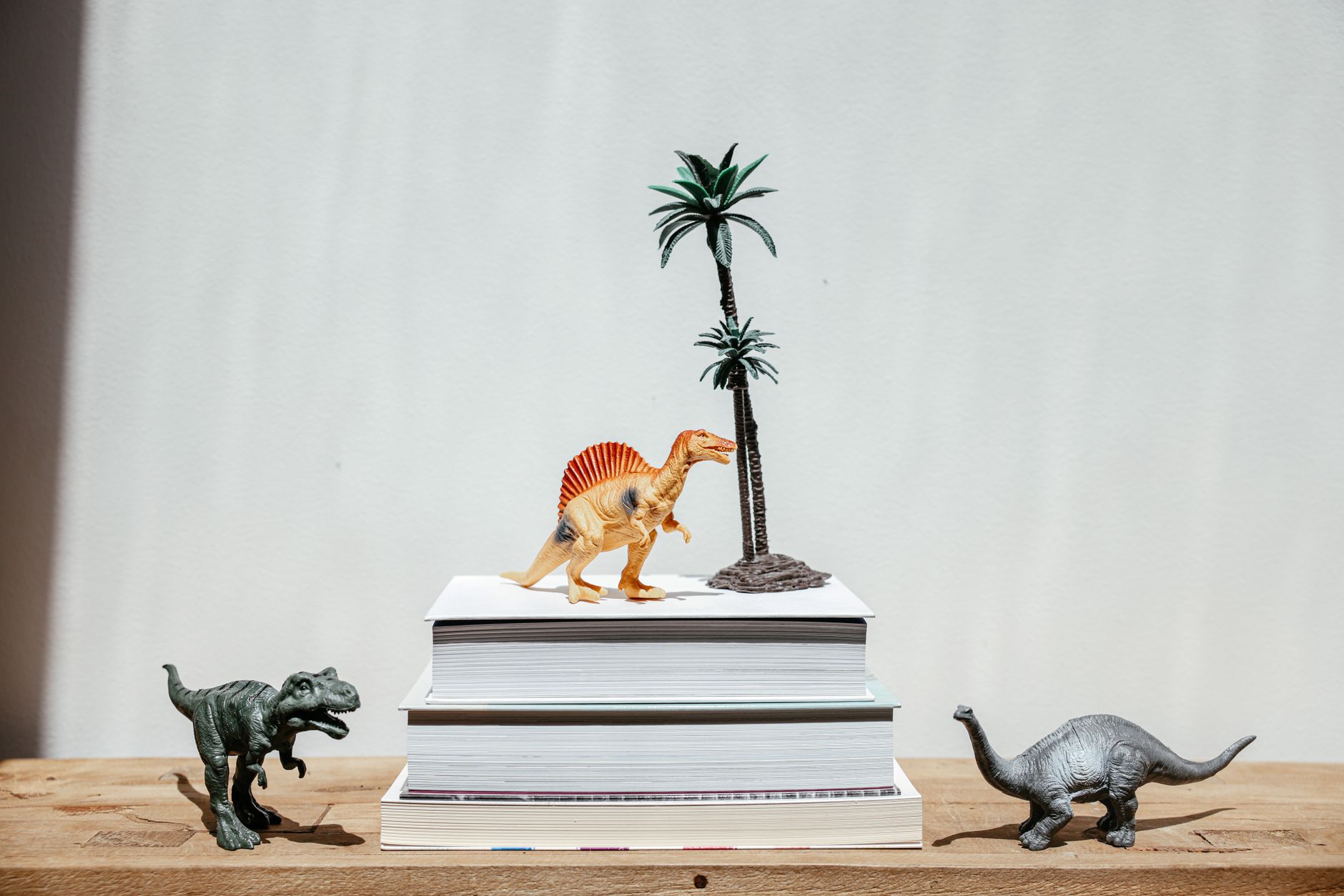 Miniature dinosaur toys on stacked books with palm trees, set on a wooden table. — free online jigsaw puzzle on JigsawPuzzlesFreeOnline.org