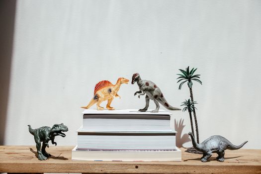 Vibrant dinosaur toys arranged on stacks of books against a plain background, creating a playful scene., free online jigsaw puzzle on JigsawPuzzlesFreeOnline.org