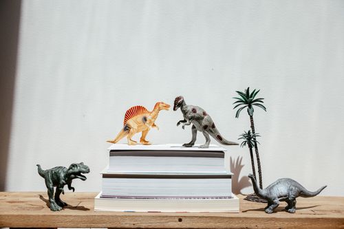 Free Vibrant dinosaur toys arranged on stacks of books against a plain background, creating a playful scene. Stock Photo