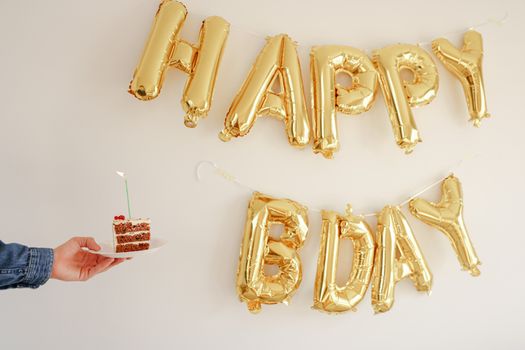 A slice of birthday cake with candle and gold 'Happy Bday' balloons on a white background.