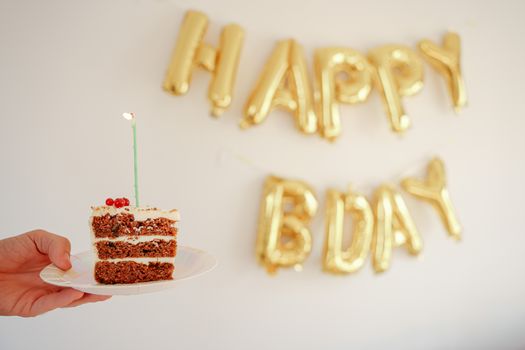 A slice of cake with a candle, held against a backdrop of gold 'Happy Bday' balloons.