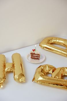Slice of cake with candle amidst gold balloon letters, perfect for birthday celebrations.