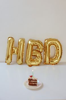 Slice of cake with lit candle and gold HBD balloons in background, perfect for celebrations.