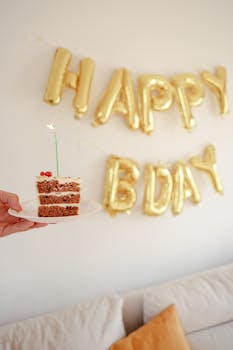 A slice of birthday cake with candle in front of gold balloons spelling 'HAPPY BDAY'.