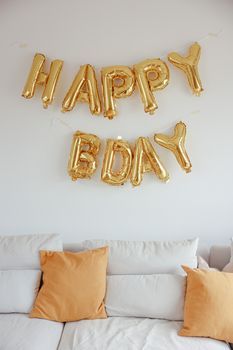 Elegant birthday setup with gold foil balloons spelling 'Happy Bday' above a cozy sofa.