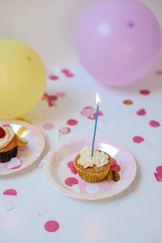 Cupcake with lit candle among balloons and confetti on a festive birthday table.