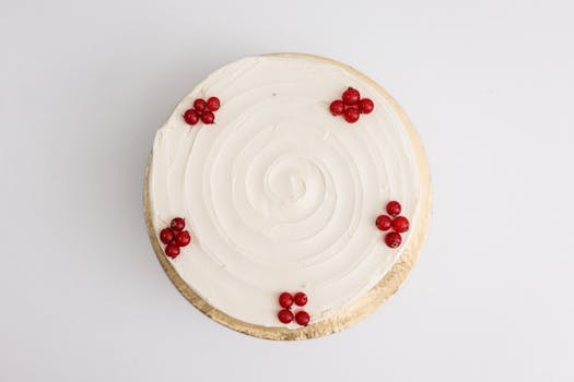 A beautifully decorated cake with white frosting and red berries, viewed from above on a white surface.