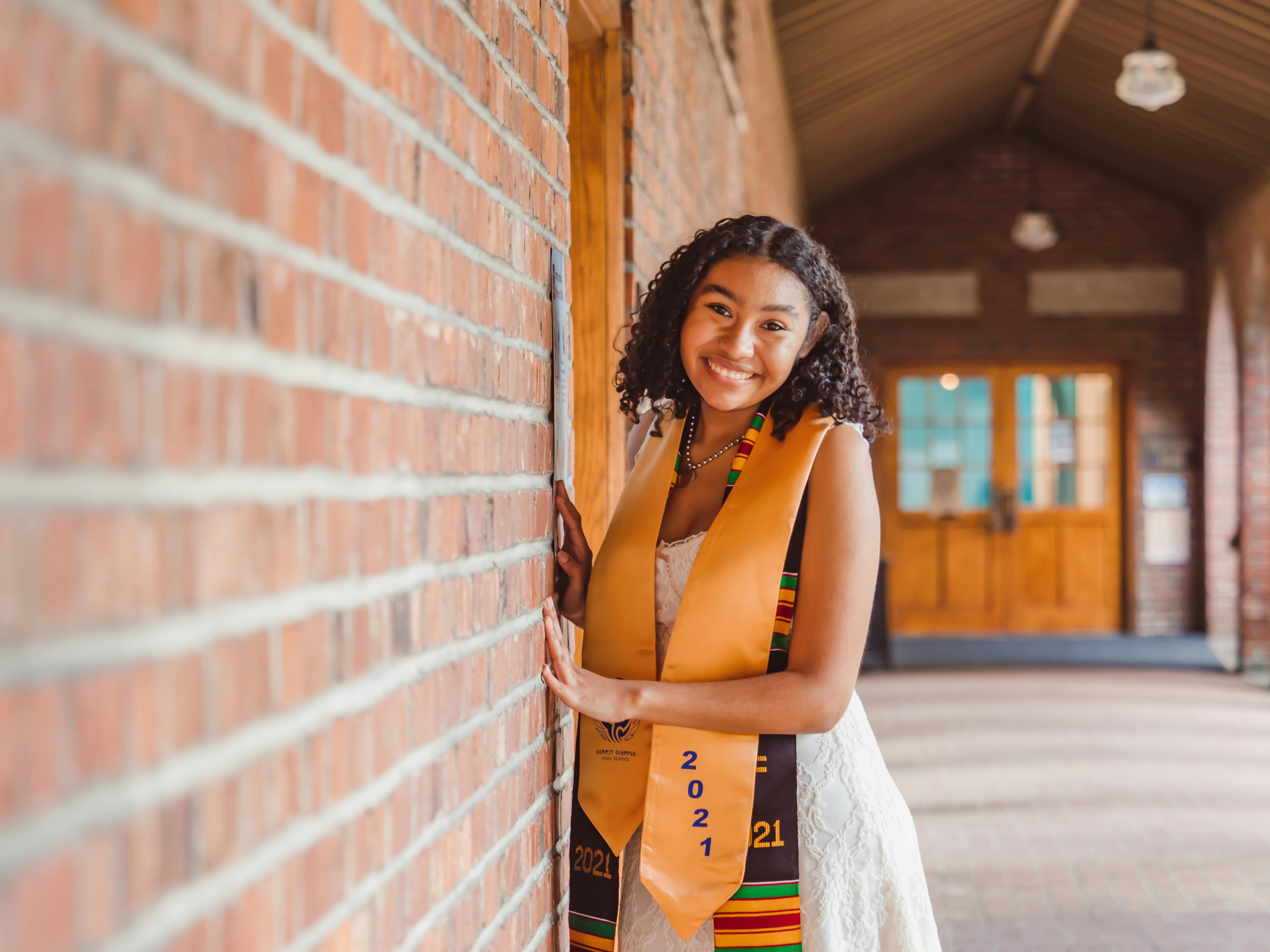 Smiling graduate posing in a brick corridor, wearing a sash.