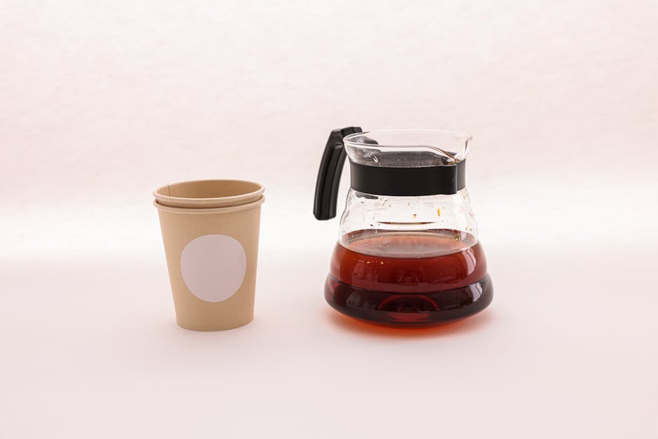 Glass coffee pot with hot beverage next to an empty paper cup on a clean white surface.