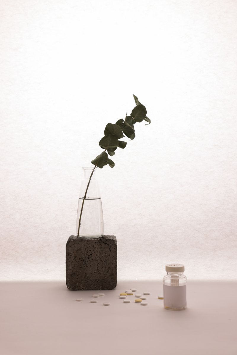 Eucalyptus branch in a vase next to a medicine bottle with pills scattered on a neutral background.