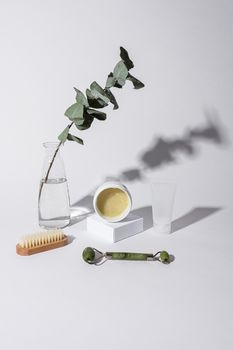 Minimalist skincare arrangement with jade roller, brush, and eucalyptus branch on white backdrop.