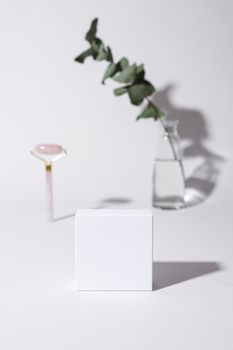 A minimalist studio setup featuring a white cube mockup, a vase with eucalyptus, and a facial roller on a white background.