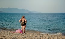 一名女子身着泳衣，站在宁静的海滩上，凝望着平静的大海和远处的山脉