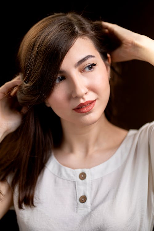 Free Young tender woman in white clothes with makeup touching brown hair while looking away in shiny light Stock Photo