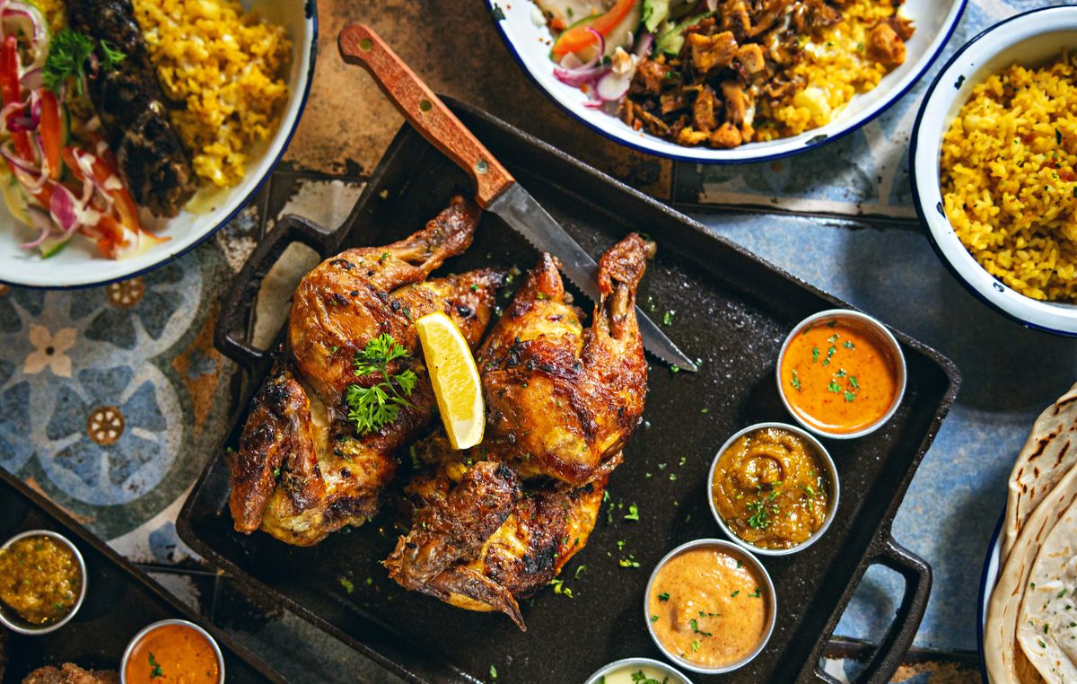 Grilled spatchcock chicken halves with lemon and parsley on black tray with sauces, yellow rice, flatbread