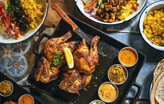 Grilled spatchcock chicken halves with lemon and parsley on black tray with sauces, yellow rice, flatbread