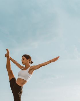 Woman practicing yoga outdoors, striking a pose against a clear blue sky. Perfect for promoting fitness and healthy lifestyle.