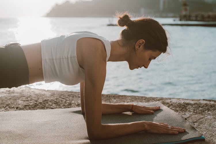 Woman In White Top Doing Plank On Elbows On Yoga Mat Beside The Sea