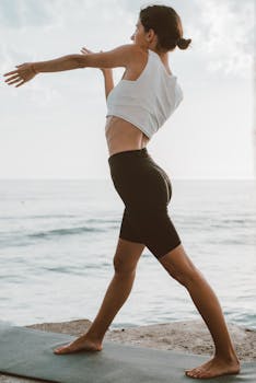 A woman in a white top and black shorts performs yoga by the seaside, enjoying a summer day.