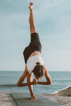 A woman practices standing split pose on a mat by the calm sea, showcasing balance and flexibility.