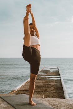 A woman practicing yoga on a pier with the ocean in the background, promoting relaxation and fitness.
