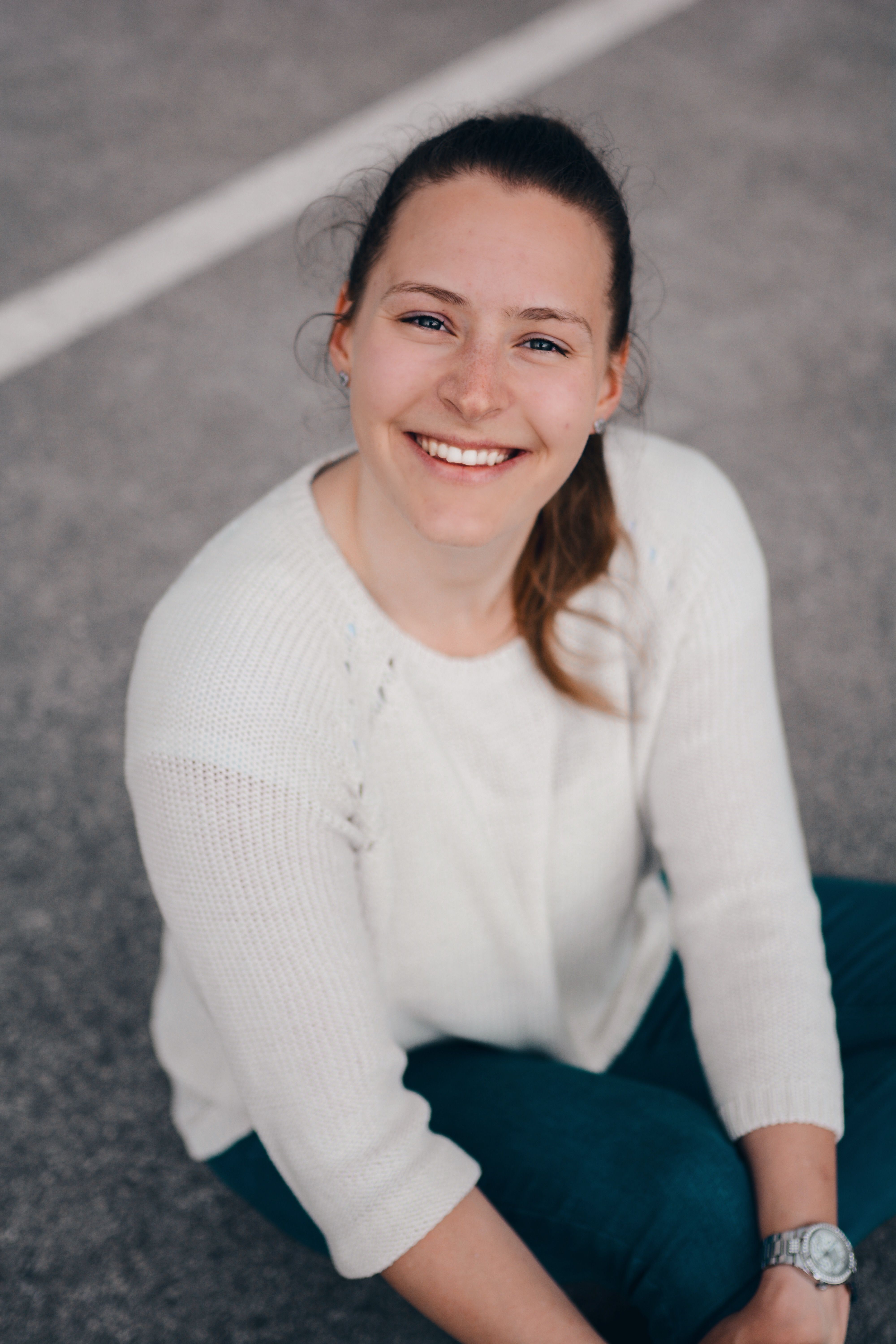 Portrait of a smiling woman in casual clothes sitting on a paved surface outdoors.