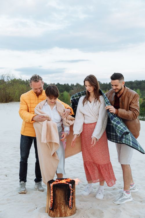 Free Four adults gathered around a bonfire on a sandy beach, wrapped in blankets, sharing warmth and togetherness. Stock Photo
